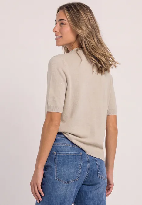 Frau in beigem Kurzarm-Pullover und blauen Jeans, seitlich mit Blick nach links vor hellem Hintergrund.