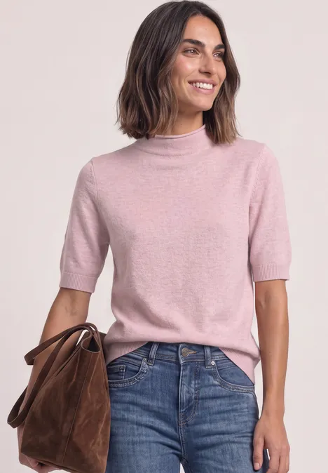 Frau mit rosa Kurzarm-Rollkragenpullover, Jeans und brauner Wildledertasche lächelt.