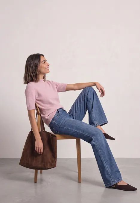 Frau sitzt seitlich auf Stuhl, rosa Oberteil, blaue Jeans, braune Schuhe, braunen Mantel an Lehne.