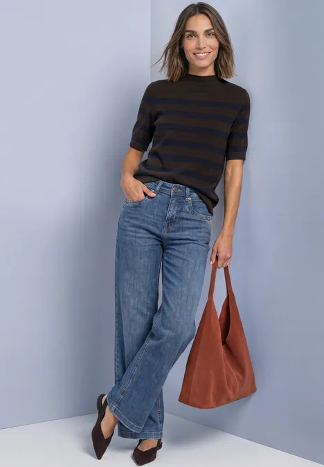 Eine Frau lächelt in gestreiftem Shirt, weiten Jeans und flachen Schuhen, hält eine braune Tasche vor hellblauer Wand.