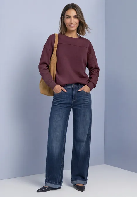 Sweatshirt mit Rundhals und Zierdetail rustic rouge Frau in lila Pullover, weiten Jeans und Handtasche lächelnd vor neutralem Hintergrund.
