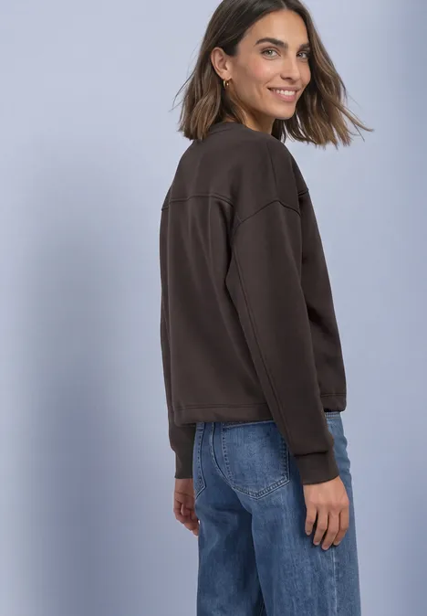 Sweat-shirt à col rond avec détails décoratifs black coffee Eine lächelnde Frau in braunem Pullover und Jeans, schaut über die Schulter vor hellem Hintergrund.