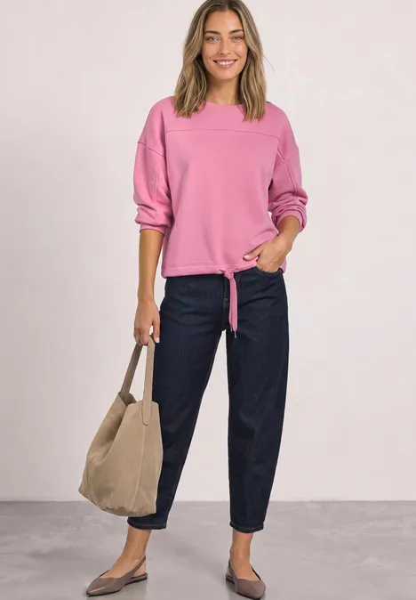 Frau in pinkem Pullover, dunklen Jeans und beiger Tasche steht lässig vor hellgrauem Hintergrund.