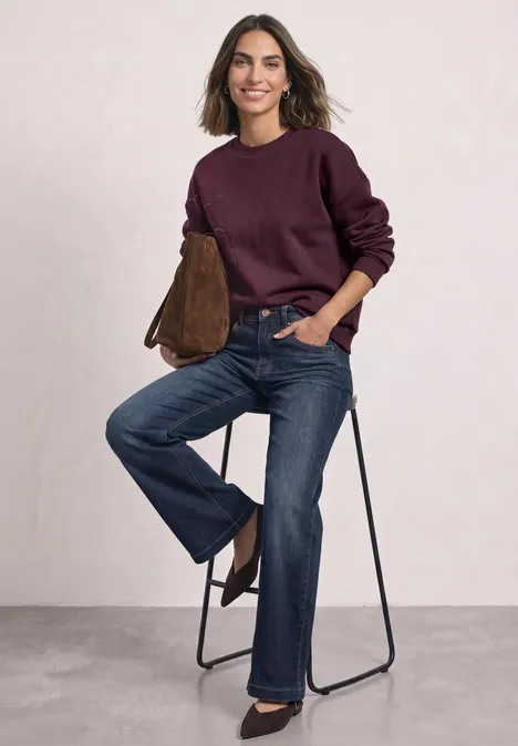 Sweatshirt met Loose Fit-pasvorm en folieprint rustic rouge Frau in Jeans und weinrotem Pullover sitzt lässig auf einem Hocker, hält eine braune Tasche.