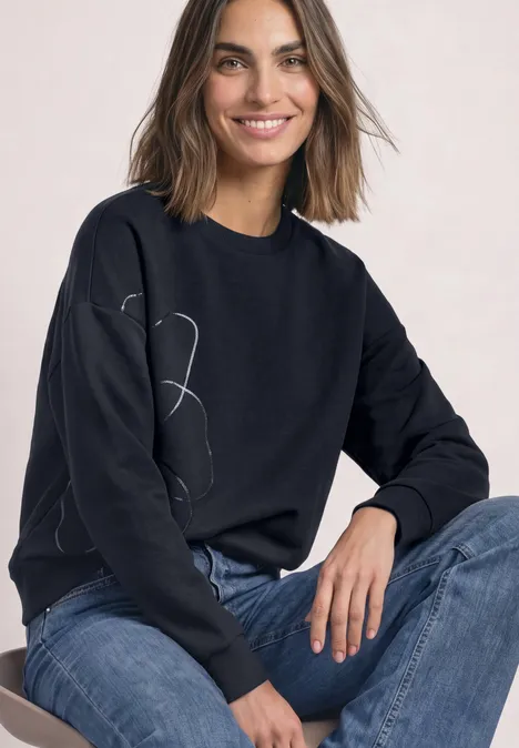 Sweatshirt met Loose Fit-pasvorm en folieprint indigo night Frau mit schwarzem Sweatshirt mit Linienzeichnung und blauen Jeans, lächelt in die Kamera.