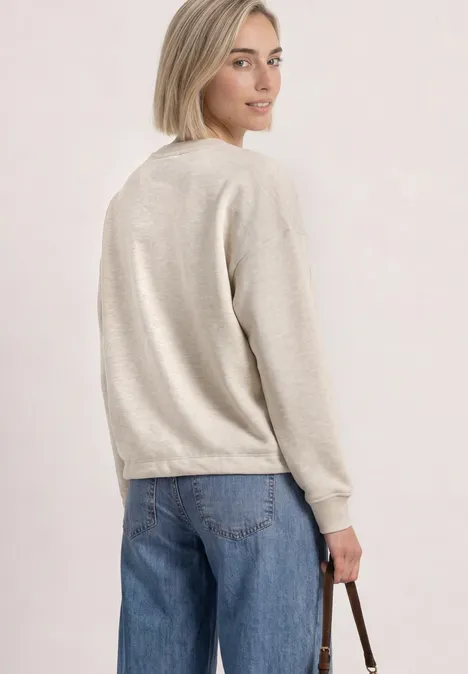 Frau in beige Pullover und Jeans, schaut über die Schulter und hält eine Tasche in der Hand.