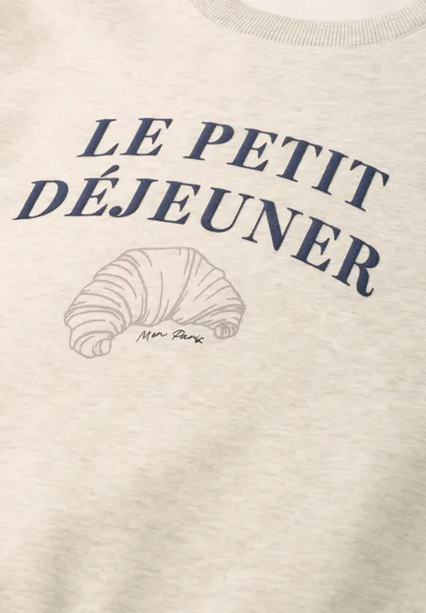 Ein cremefarbenes T-Shirt mit der Aufschrift 'Le Petit Déjeuner' und einer Croissant-Skizze.