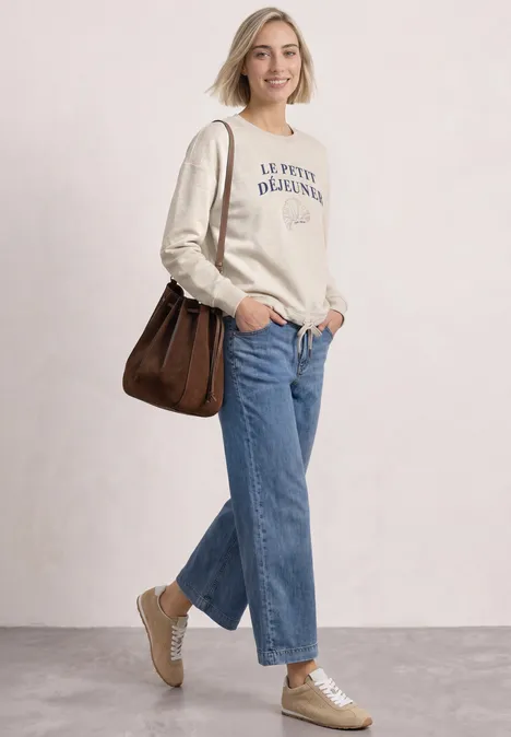 Junge Frau in legerem Outfit mit Creme-Sweatshirt, blauen Jeans, beigen Sneakern und brauner Tasche, lächelnd.