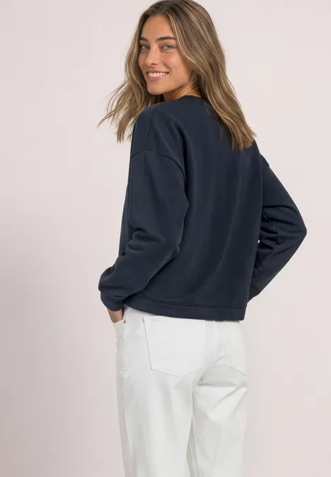 Frau mit dunklem Sweatshirt und weißer Hose blickt über die Schulter und lächelt.