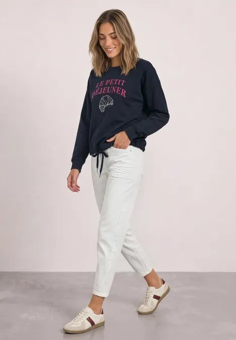 Frau im blauen Sweatshirt, weißen Jeans und Sandalen, lächelnd in lässiger Pose.