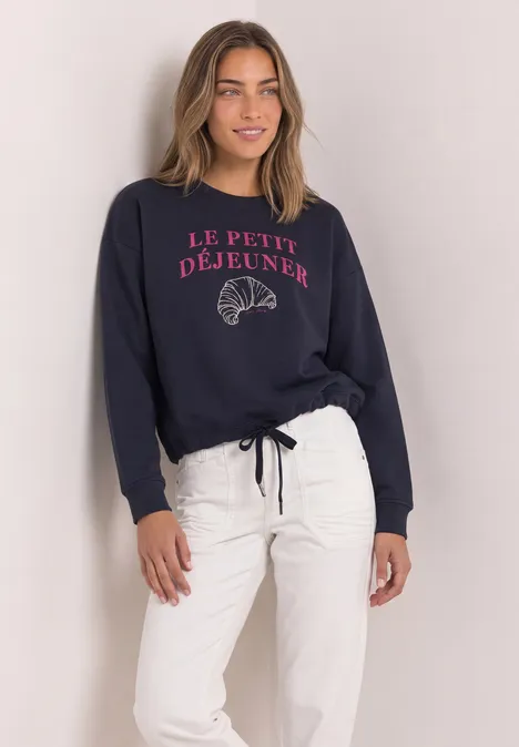 Eine Frau trägt ein dunkelblaues Sweatshirt mit 'Le Petit Déjeuner'-Aufdruck und weiße Hose.