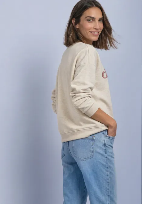 Lächelnde Frau in beigem Pullover und blauer Jeans, seitlich stehend, Hände in den Taschen.