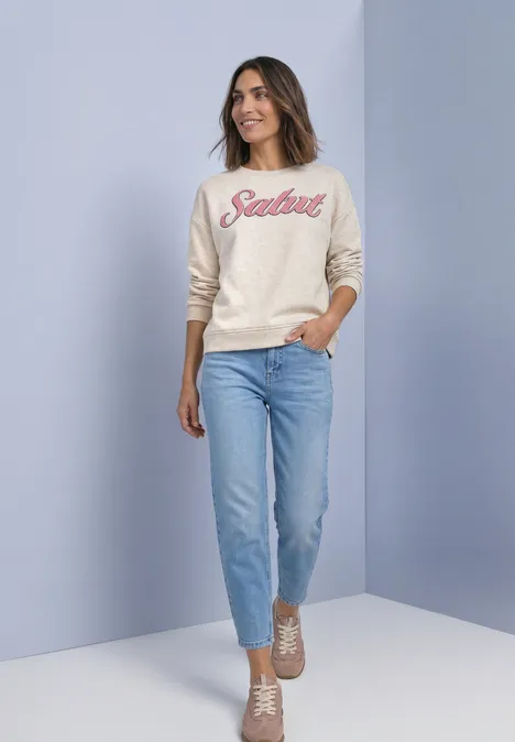 Eine Frau in Jeans, hellem Pullover mit 'Salut' Schriftzug und beigen Sneakern vor pastellfarbenem Hintergrund.
