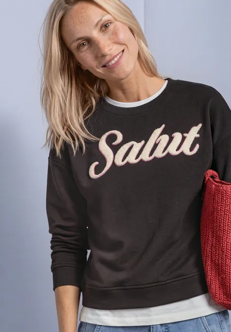 Eine lächelnde blonde Frau trägt ein schwarzes Sweatshirt mit dem Schriftzug "Salut" und hält eine rote Tasche.