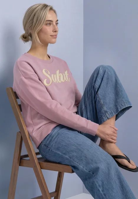 Junge Frau sitzt seitlich auf einem Klappstuhl, trägt rosa Sweatshirt mit „Salut“, weite Jeans und Flip-Flops.