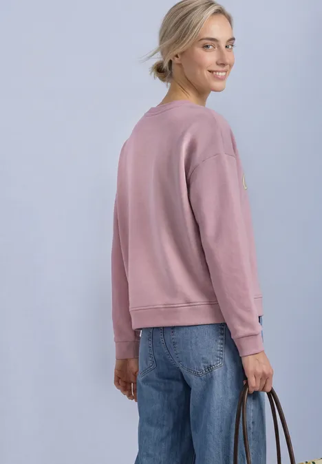 Eine Frau im rosa Pullover und blauen Jeans blickt über die Schulter und hält eine Tasche in der Hand.