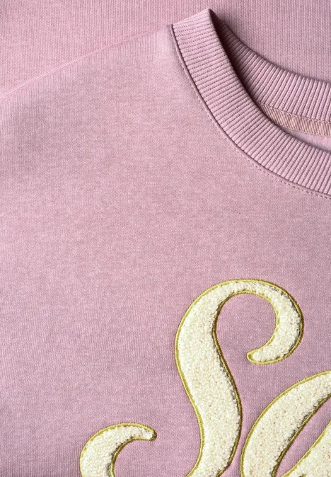 Rosafarbener Pullover mit goldglitzerndem Buchstaben-Detail, Nahaufnahme.