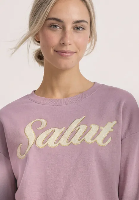 Junge Frau im rosa Sweatshirt mit goldener 'Salut'-Aufschrift lächelt dezent.