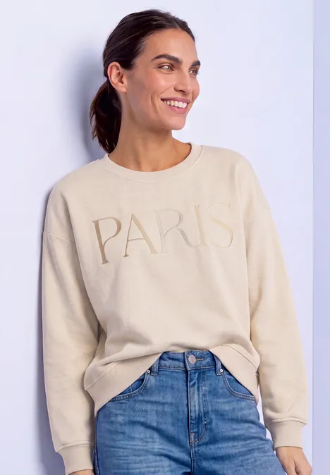 Frau in beigem Pullover mit 'PARIS'-Schriftzug und Jeans lächelt seitlich.