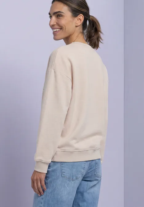 Junge Frau im beigen Pullover und Jeans, seitlich stehend vor lila Hintergrund, lächelnd.