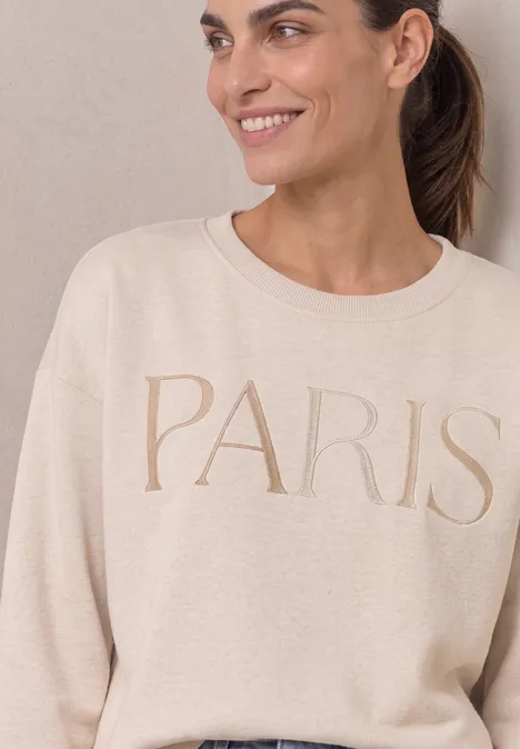 Eine lächelnde Frau in einem beigen Pullover mit der Aufschrift PARIS.