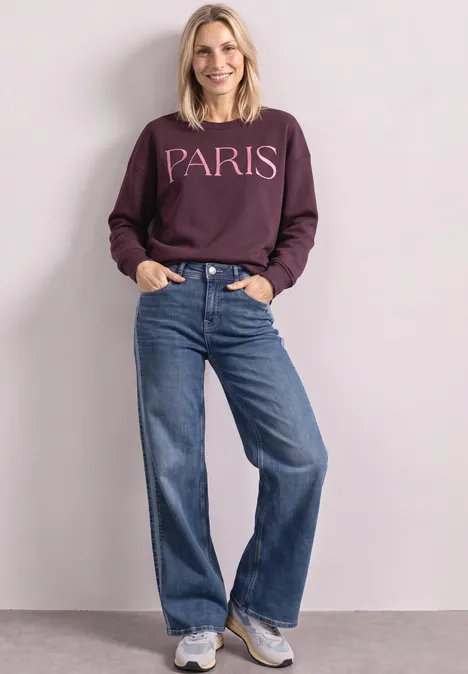 Jeune femme souriante en sweat violet 'PARIS', jean large et baskets, posant devant un mur clair.