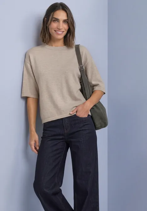 Frau mit beige kurzem Pullover, dunklen Jeans und grüner Umhängetasche vor hellem Hintergrund, lächelnd.