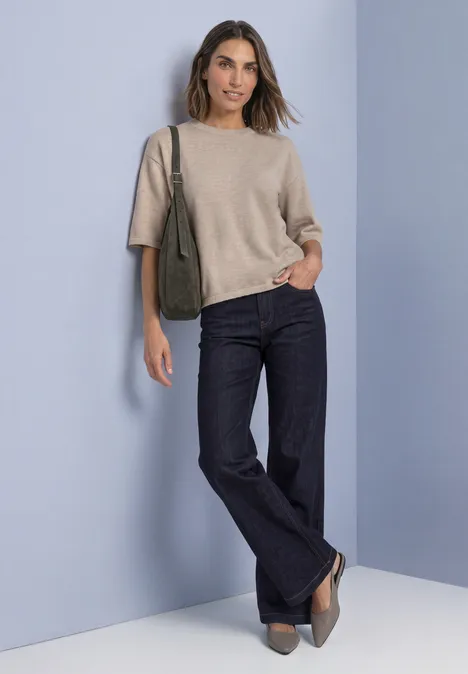 Eine Frau trägt einen beigen Pullover, dunkle Jeans und graue Schuhe vor hellem Hintergrund.