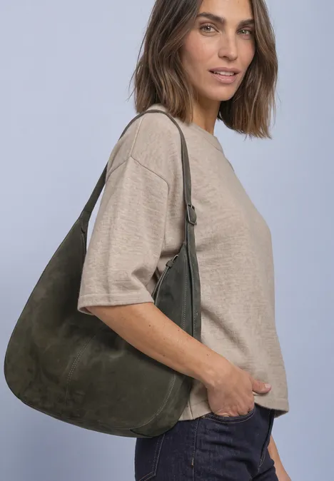 Frau mit beiger Bluse und dunkler Jeans trägt eine große grüne Wildleder-Schultertasche und lächelt leicht.