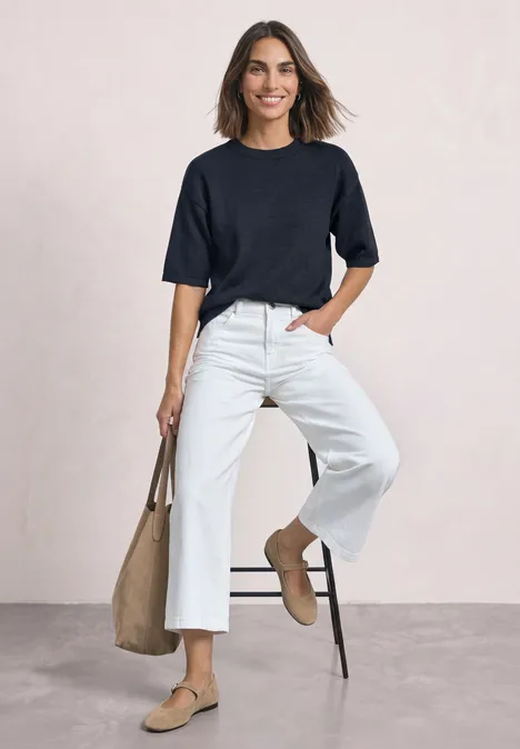 3/4-Arm Pullover mit Rundhals darkest blue mel. Frau in schwarzem Kurzarmshirt, weißer Culotte, beigen Loafers und Tasche lehnt lächelnd an Stuhl.