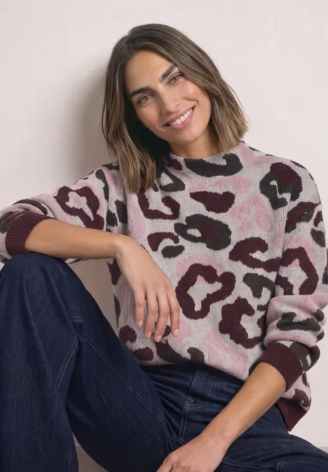 Trui met col en luipaardpatroon pure beige Eine lächelnde Frau sitzt entspannt, trägt einen rosa-braunen Pullover mit Muster und eine blaue Jeans.