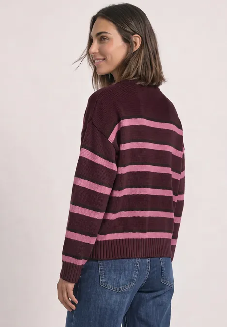Frau mit schulterlangem Haar trägt burgunderfarbenen Pullover mit pinken Streifen und Jeans, Blick über die Schulter.