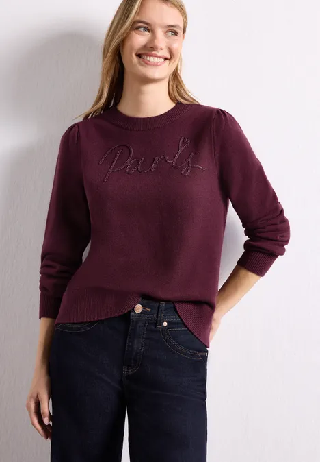 Femme souriante portant un pull prune orné d'un motif brodé et un jean foncé, mains dans les poches.