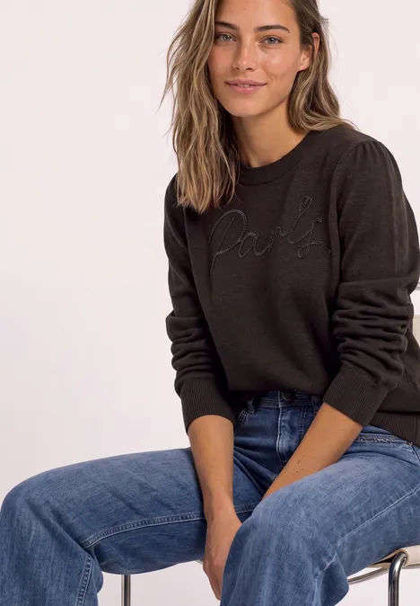 Pullover mit Emboidery Wording black coffee Frau sitzt auf einem Stuhl, trägt schwarzen 'Paris'-Pullover und blaue Jeans, lächelt entspannt.