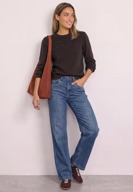 Pullover mit Emboidery Wording black coffee Eine Frau in dunklem Pullover mit Stofftasche trägt lässige Jeans und braune Schuhe vor heller Wand.