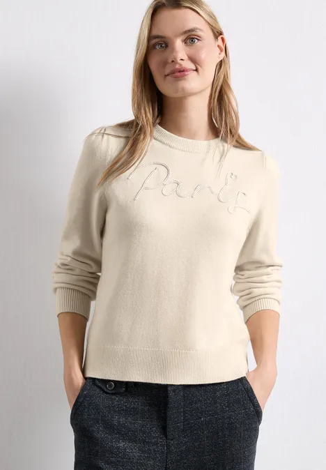 Femme portant un pull crème 'Paris' et un pantalon à carreaux gris, mains dans les poches.