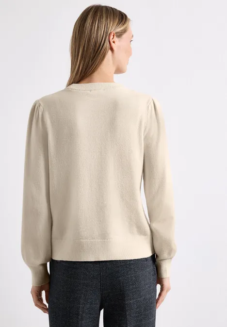 Femme de dos en pull beige à manches longues et pantalon gris sur fond blanc.
