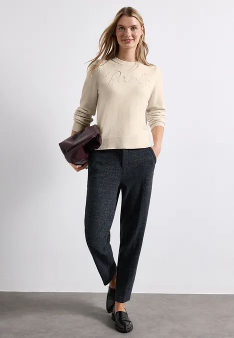 Une femme porte un pull crème et un pantalon gris, tenant une veste pliée, debout sur fond neutre.
