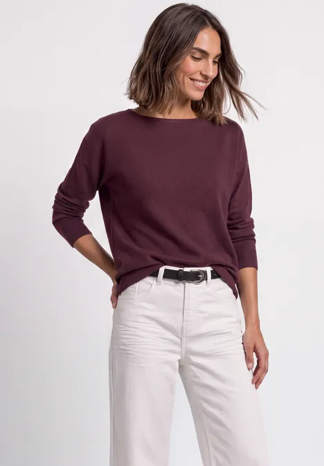 Dame trägt bordeauxroten Pullover und helle Jeans, lächelnd in entspannter Pose.
