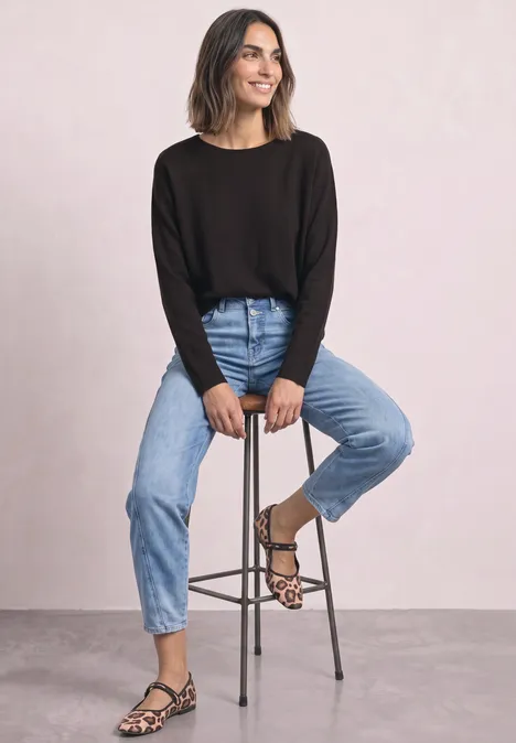 Vrouw lacht, zit op kruk, draagt zwarte trui, blauwe jeans en luipaardprint sandalen.