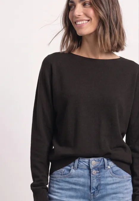 Vrouw met donkerbruine trui en high-waist jeans, glimlacht zijwaarts.