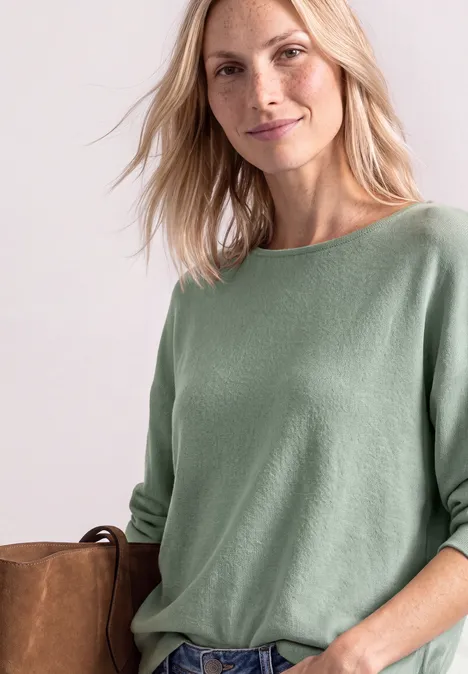 Dolman-trui met boothals eucalyptus green mel. Een blonde vrouw draagt een lichtgroene trui en houdt een bruine suède tas vast.