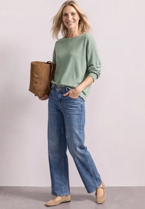 Dolman-trui met boothals eucalyptus green mel. Vrouw in groene blouse en spijkerbroek met een bruine tas en beige schoenen, voor een lichte achtergrond.