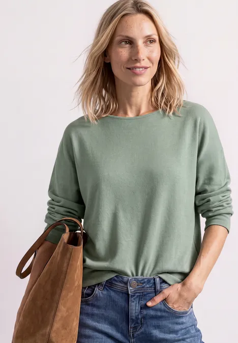 Dolman-trui met boothals eucalyptus green mel. Vrouw glimlacht in groen trui en spijkerbroek, met bruine tas voor een witte achtergrond.