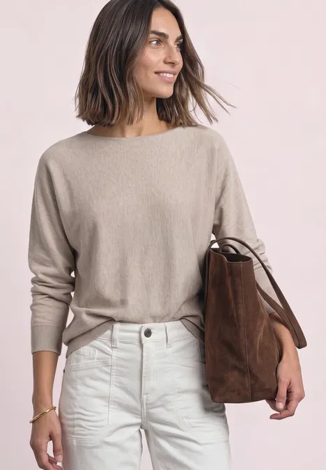 Vrouw met halflang haar in beige trui en witte jeans, met een bruine suède tas over de schouder.