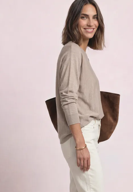 Vrouw in beige trui en witte broek, met grote bruine tas en een glimlach.