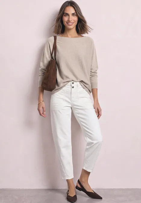 Een vrouw draagt een beige trui, een witte broek, een bruine schoudertas en donkere ballerina’s voor een lichte muur.