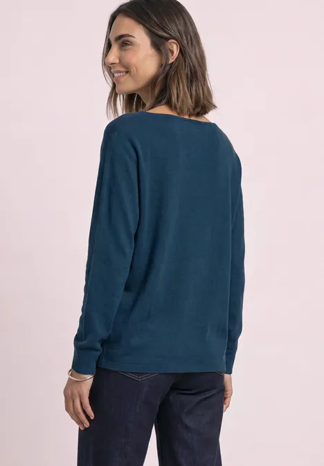 Dolman Pullover mit Boat Neck blue petrol Frau im dunkelblauen Pullover und Jeans, seitlich stehend, Blick über die Schulter vor rosa Hintergrund.