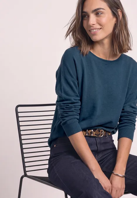 Dolman Pullover mit Boat Neck blue petrol Frau sitzt lächelnd auf einem Drahtstuhl, trägt blauen Pullover und dunkle Jeans.
