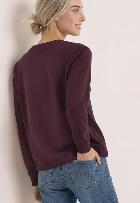 Frau in bordeauxrotem Pullover und Jeans lehnt seitlich an einer Wand.